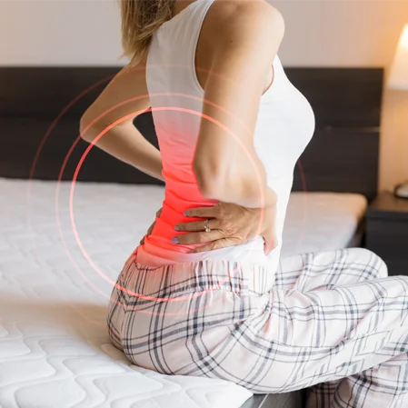 A woman sitting on the edge of her bed and holding her back in pain